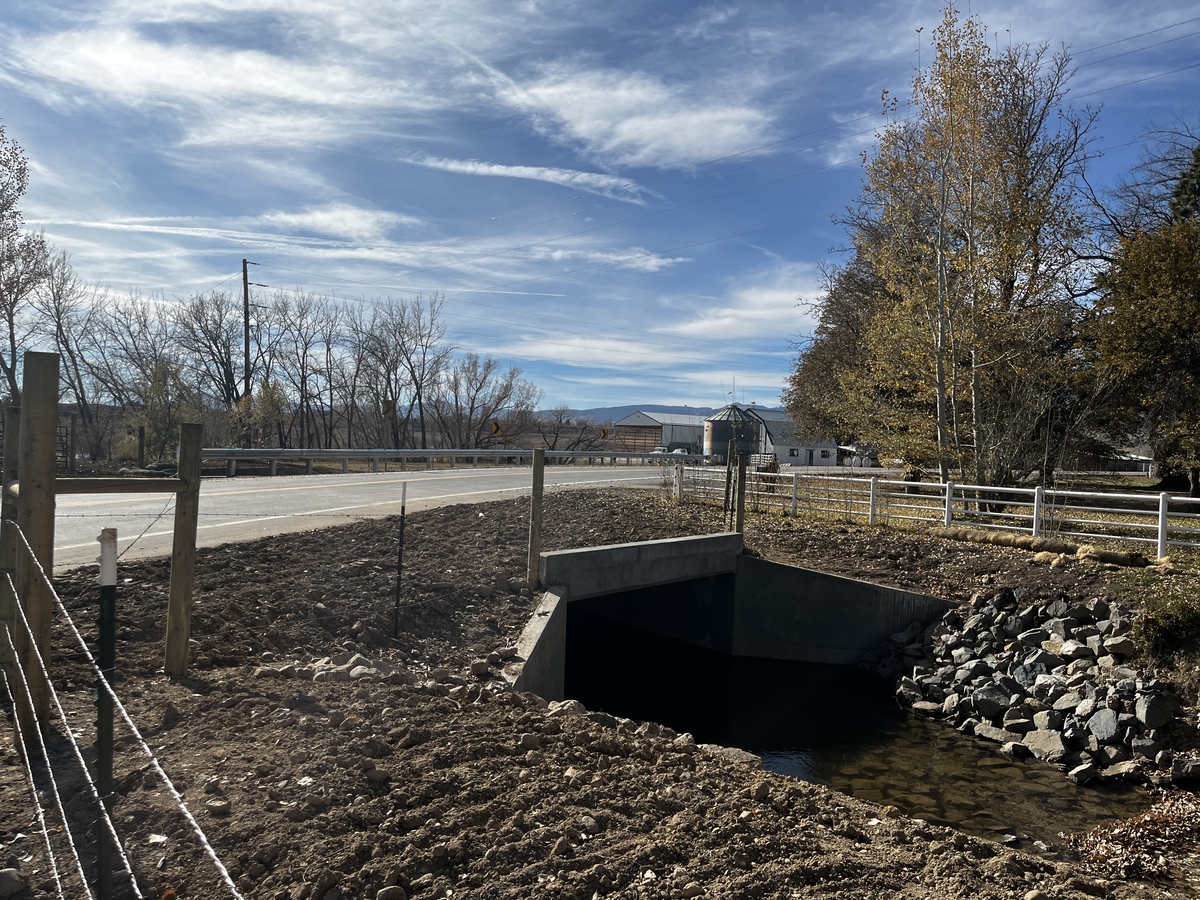 Road crossing over a drainage culvert, with a small stream flowing beneath the roadway beside a fenced rural roadside and autumn trees.